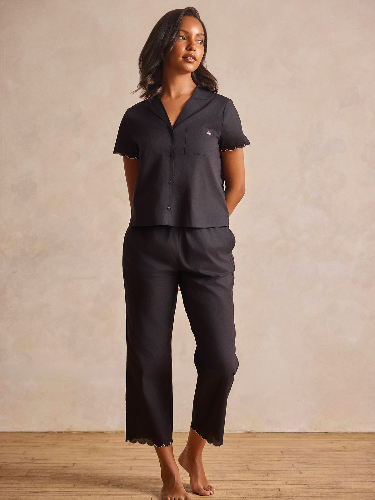A woman modeling a dark charcoal gray two-piece pajama set with a short-sleeve button-front top and matching cropped pants, standing against a textured beige wall and light wood floor.