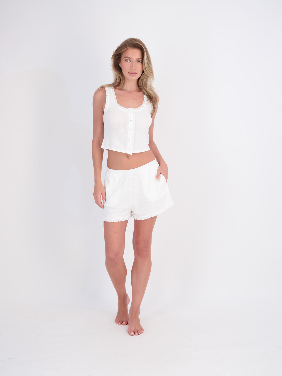 A woman modeling a white two-piece lounge set consisting of a sleeveless cropped button-front top and matching shorts, standing against a seamless white studio background.